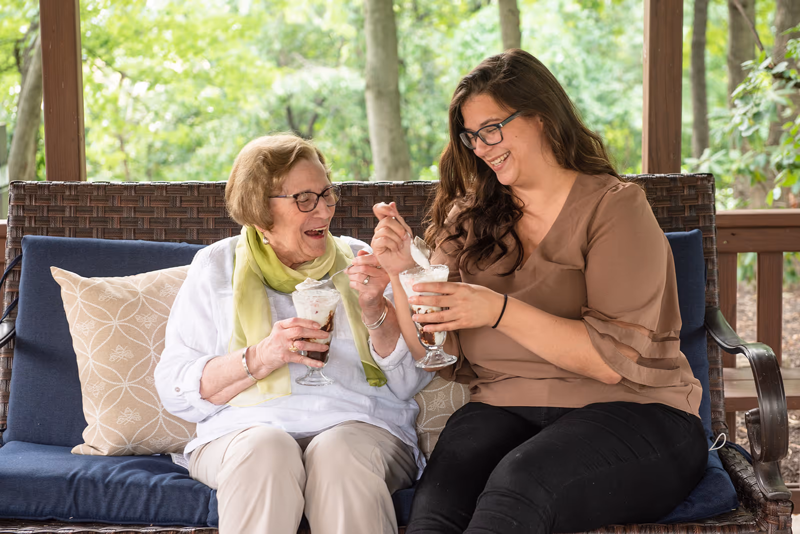 An elderly woman and a younger woman sitting together on a cushioned outdoor wicker bench, smiling and enjoying ice cream sundaes on a porch with green trees in the background.