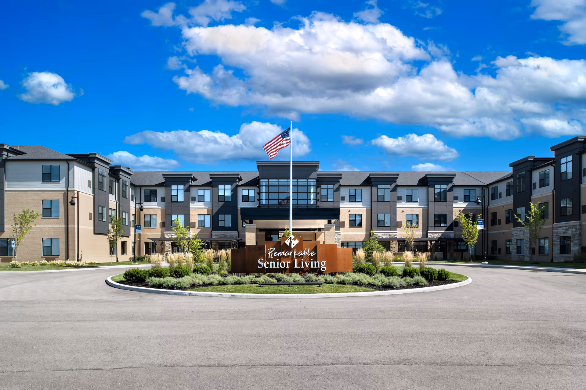 Front exterior view of a modern three-story senior living facility building under a bright blue sky with scattered clouds. In front of the building is a circular driveway with a landscaped island featuring a sign that reads 'Remarkable Senior Living' and an American flag on a flagpole.
