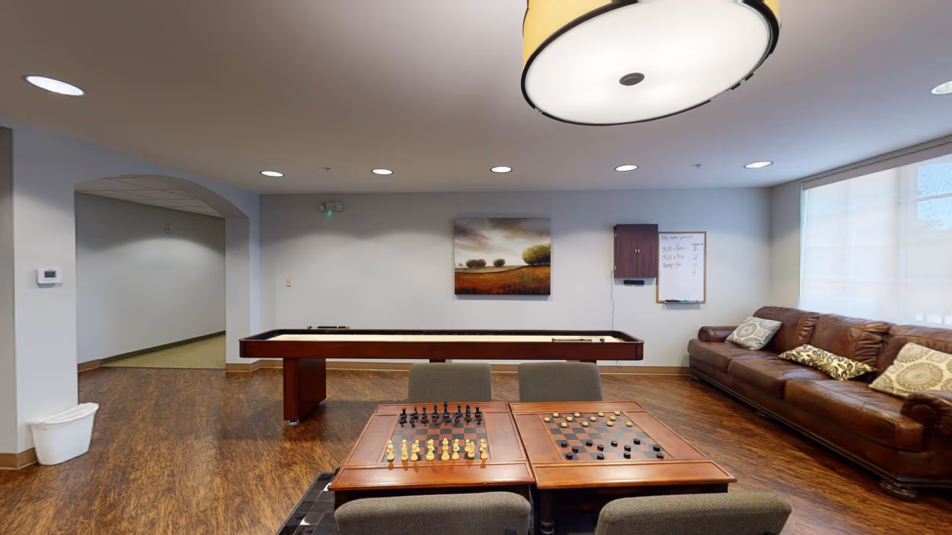A recreational room with a shuffleboard table in the center, a wooden table with chess and checkers boards in the foreground, a brown leather couch with decorative pillows along the right wall, and a landscape painting on the back wall. The room has wood flooring, recessed ceiling lights, and a large window with a translucent shade.