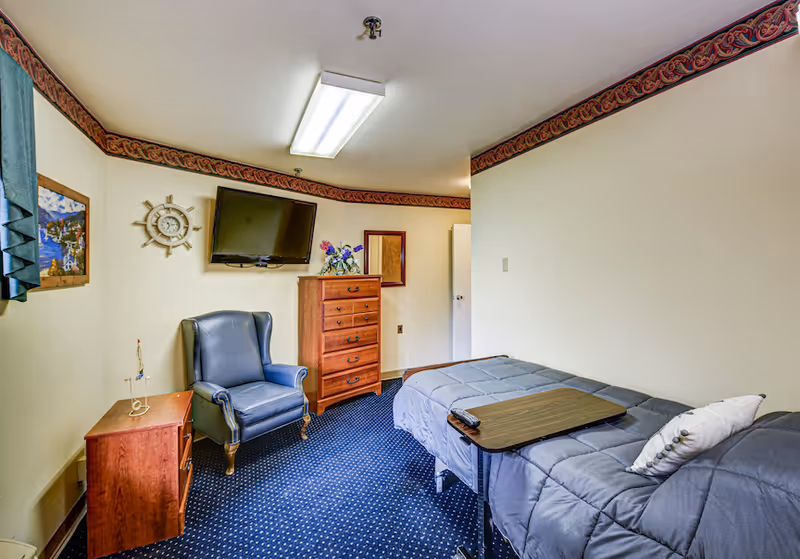 A small bedroom in a senior living facility with a single bed covered in a gray comforter and a pillow. A wooden overbed table with a TV remote is placed on the bed. The room has a blue armchair, a wooden chest of drawers with a flower arrangement on top, and a wall-mounted flat-screen TV. The walls are light-colored with a decorative border near the ceiling, and there is a framed picture and a nautical-themed clock on the wall. The floor is carpeted in blue with a dotted pattern.