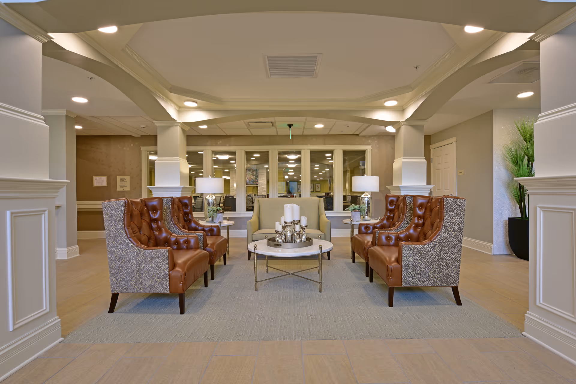 A seating area in a senior living facility with four brown leather and fabric armchairs arranged around a round coffee table with candles and decorative items. The space features soft lighting, a beige carpet, white columns, and a large window in the background showing another room.