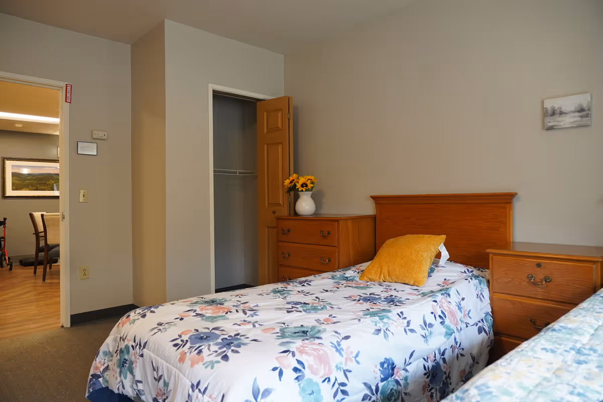 A bedroom in a senior living facility featuring a single bed with floral bedding and a yellow pillow. There is a wooden headboard, a matching wooden dresser with a vase of sunflowers, and a nightstand. An open closet door is visible, and the room has neutral-colored walls. Through the open door, a dining area with chairs and a framed picture on the wall can be seen.