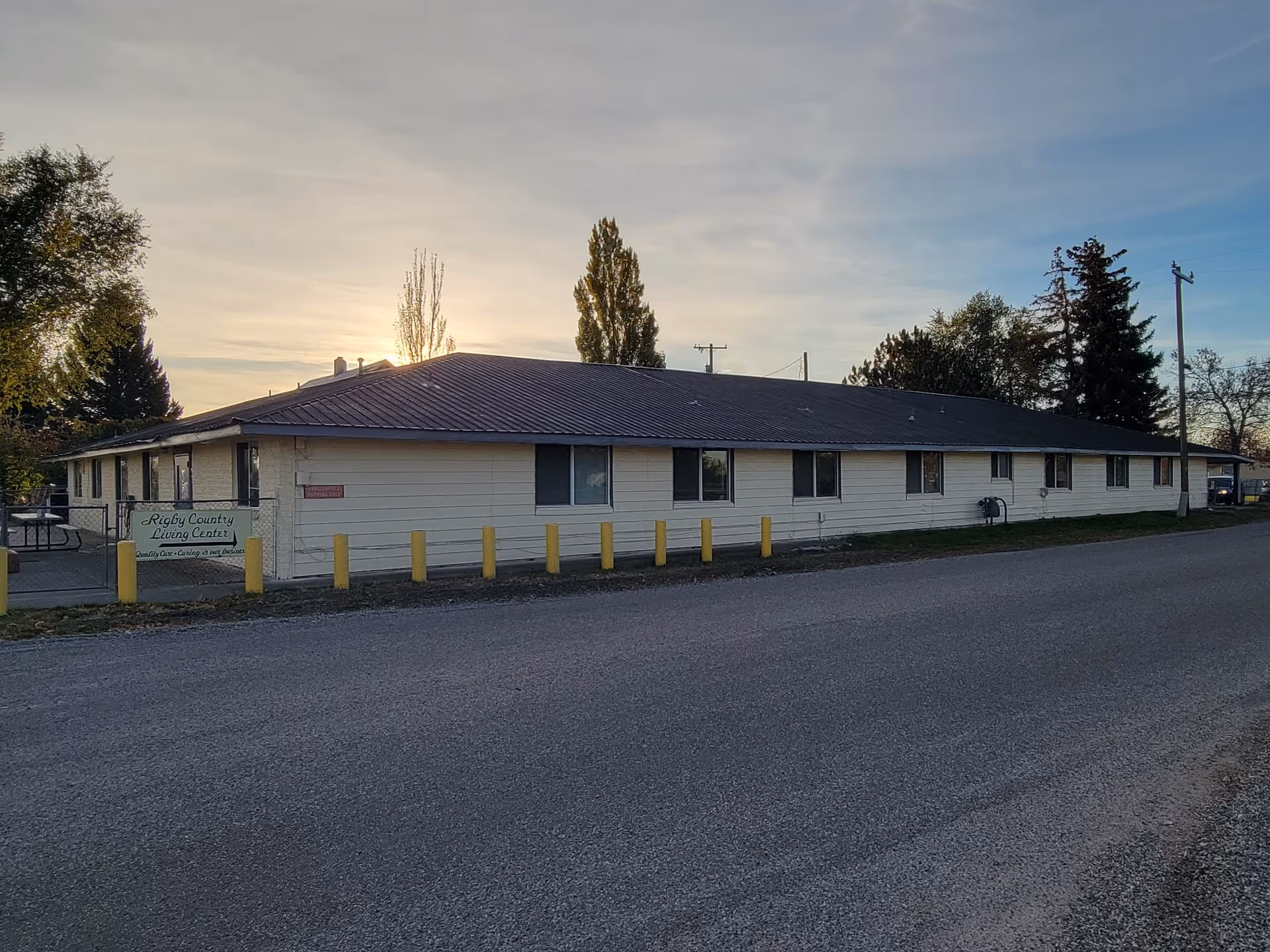 Exterior view of a single-story building with a brown roof and light-colored siding, identified as Rigby Country Living Center by a sign near the entrance. The building is surrounded by trees and is situated along a paved road during sunset.