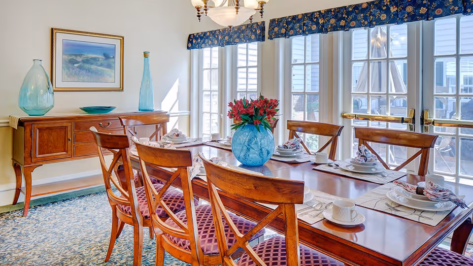 A bright dining room with a wooden dining table set for six people, featuring white plates, cups, and napkins. The table has a blue vase with red flowers as a centerpiece. Behind the table is a wooden sideboard with decorative glass vases and a framed painting on the wall. Large windows with blue floral valances let in natural light and offer a view of an outdoor patio area.