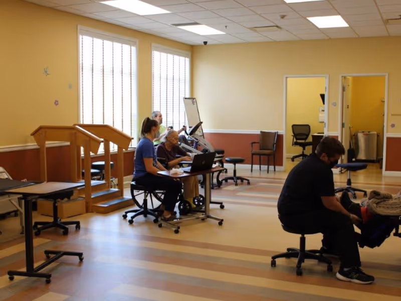 A rehabilitation room with several people engaged in therapy activities. One person is seated on a wheelchair using a laptop with assistance from a staff member, another person is using an exercise bike, and a staff member is attending to a patient lying on a therapy table. The room has large windows with blinds, beige and brown walls, and a striped floor.