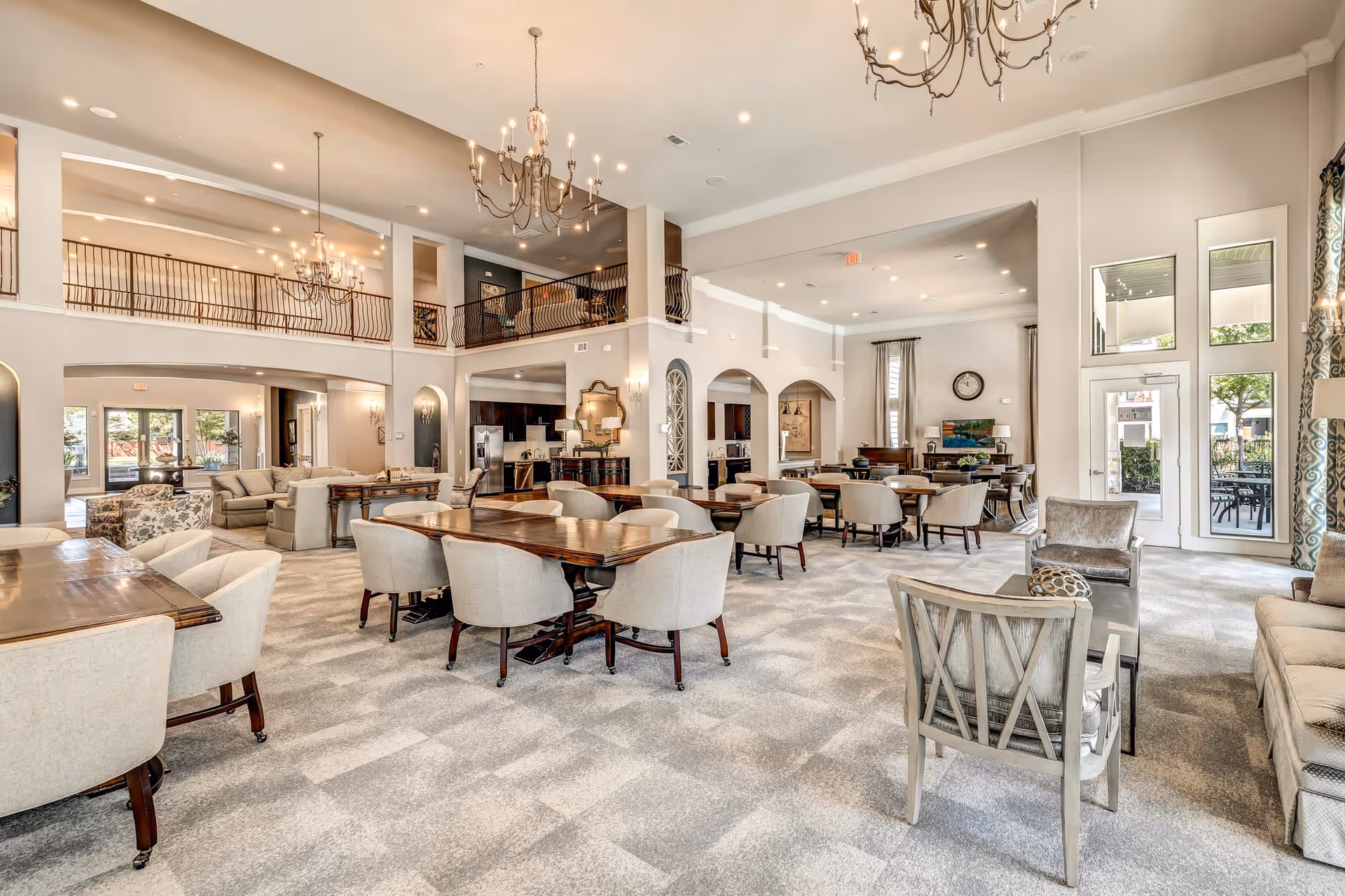 Spacious and well-lit common area in a senior living facility with multiple wooden tables and beige upholstered chairs arranged for group seating. The room features high ceilings with elegant chandeliers, a carpeted floor, large windows with patterned curtains, and a seating area with sofas and armchairs near the entrance to an outdoor patio.