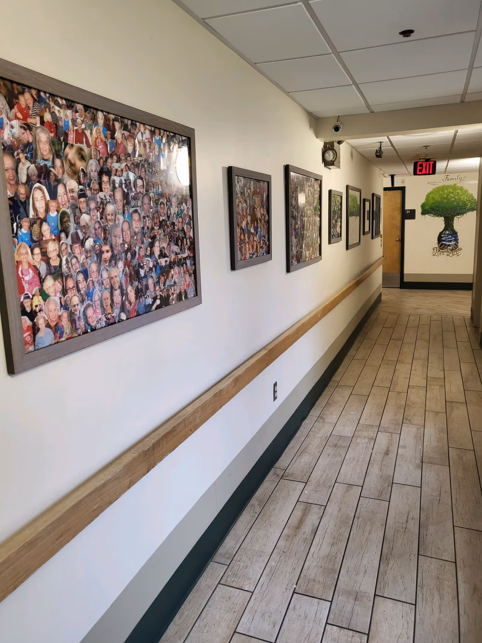 A hallway in a senior living facility with framed photo collages of elderly people on the left wall. The floor is tiled with light wood-like tiles, and there is a wooden handrail along the wall. At the end of the hallway, there is a door and a mural of a tree with the words 'Family' and 'Together' on the wall. An exit sign is visible above the door.
