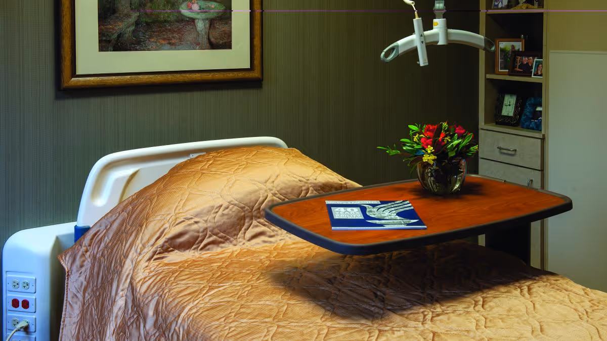 A hospital-style bed with a brown quilted blanket in a room. A wooden overbed table is positioned over the bed, holding a vase with red and yellow flowers and a blue booklet. The background includes a framed painting on the wall and a shelving unit with framed photos and drawers.