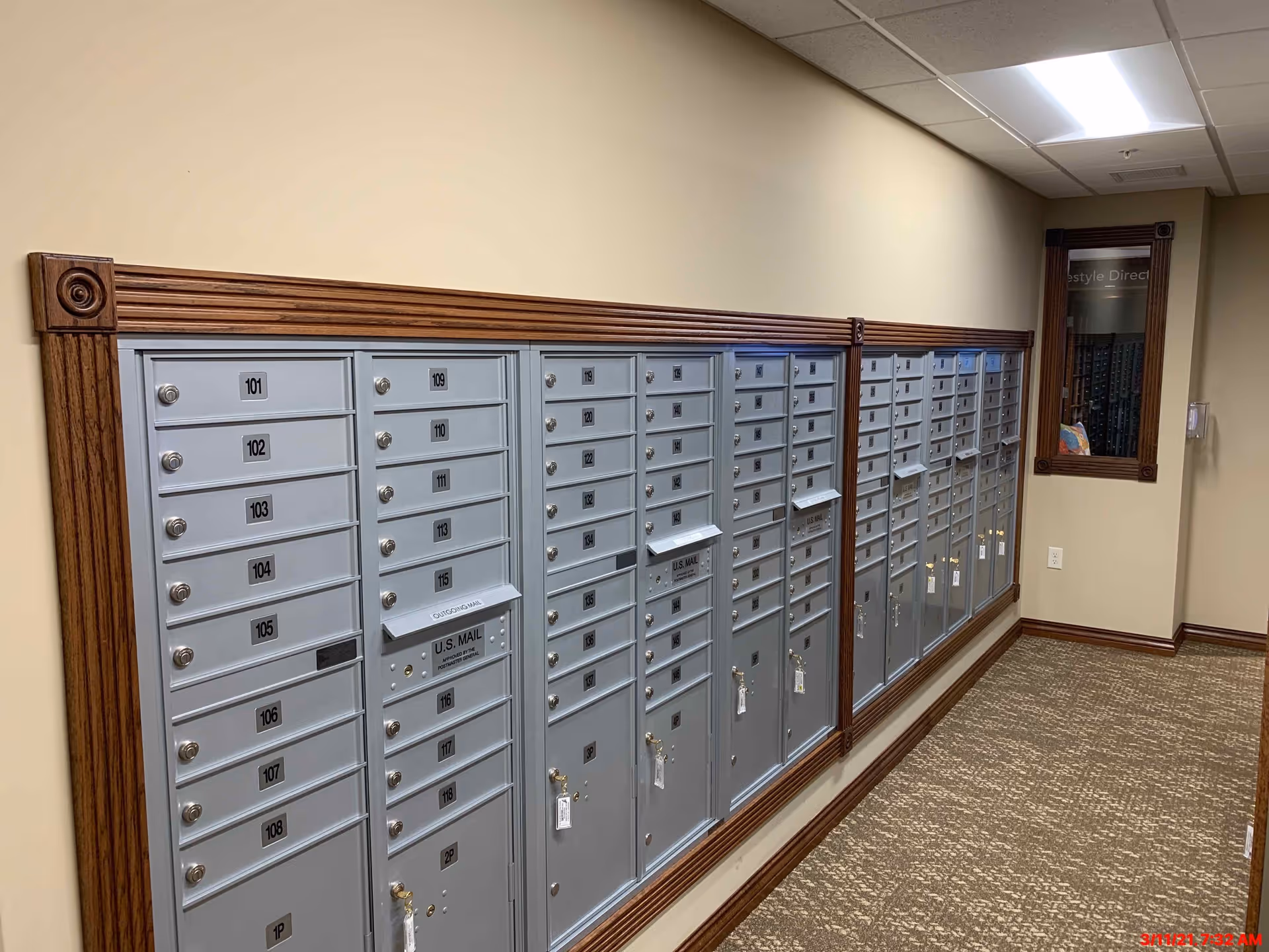 A hallway with a row of gray metal mailboxes mounted on the wall, each labeled with numbers. The wall is beige with wooden trim, and there is a carpeted floor. A mirror with a wooden frame is visible on the right side of the hallway.