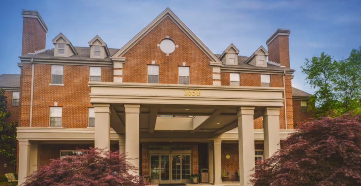 Three-story red brick building with a columned portico entrance displaying the number 1658, framed by shrubs and trees.