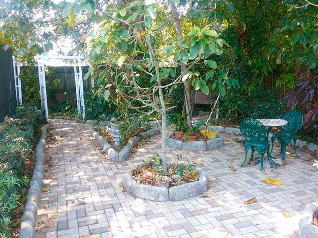 A paved outdoor garden area with a small tree planted in a circular stone border in the center. There is a green metal table with two matching chairs on the right side. The garden is surrounded by various plants and trees, and there is a white wooden archway at the far end of the paved path.