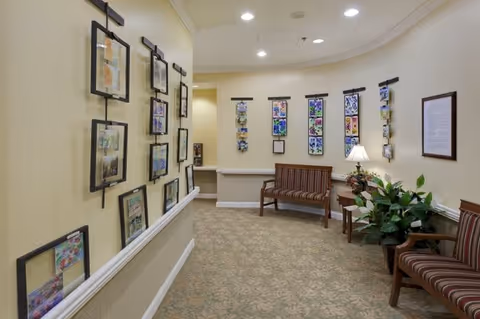 A hallway in a senior living facility with beige walls and carpeted floor. The walls are decorated with framed artwork and colorful pictures. There are two wooden benches with striped cushions along the walls, a small table with a lamp and a plant, and additional greenery in the corner. The space is softly lit with recessed ceiling lights.
