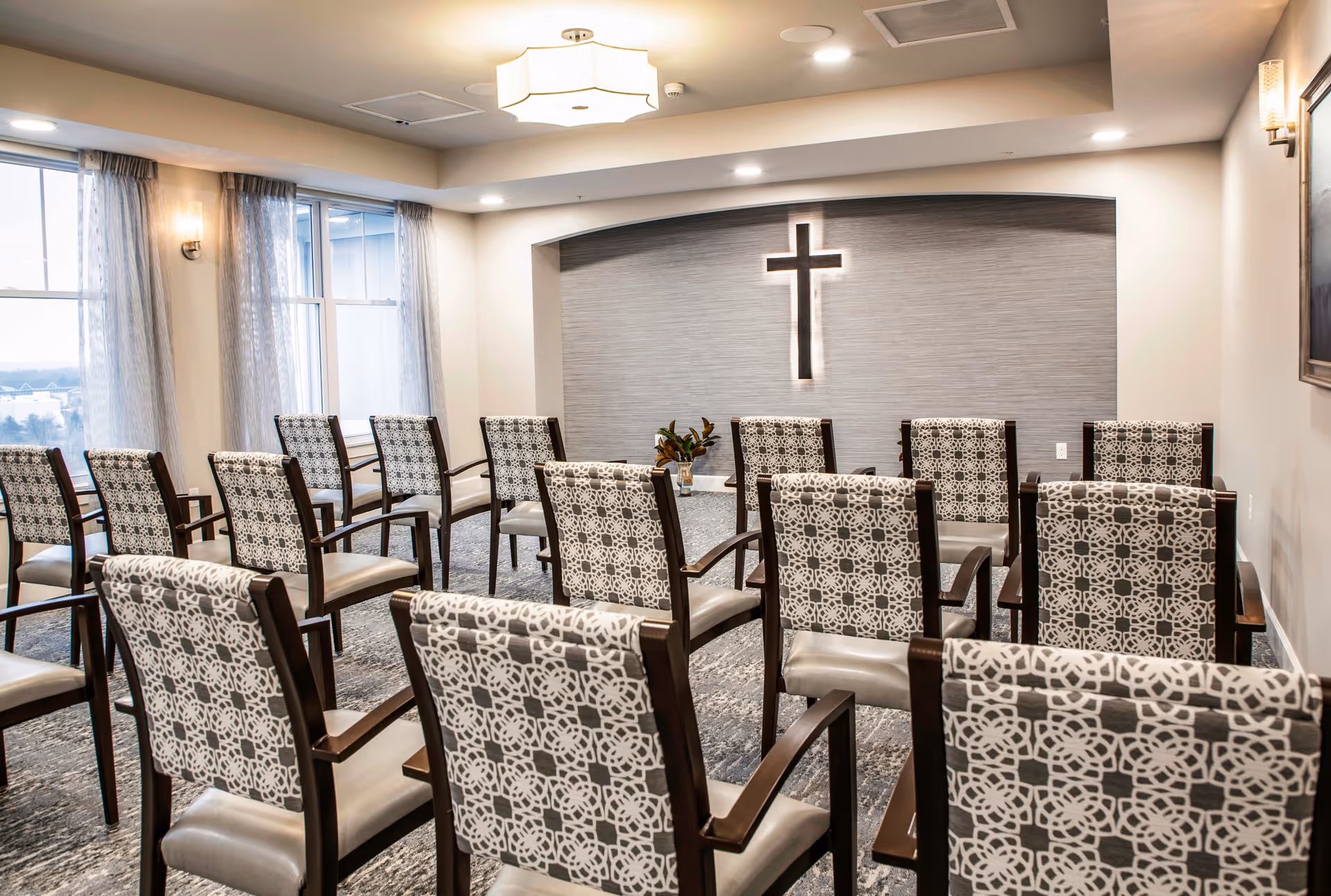 Small chapel-style meeting room with rows of patterned chairs facing an illuminated cross on the wall.