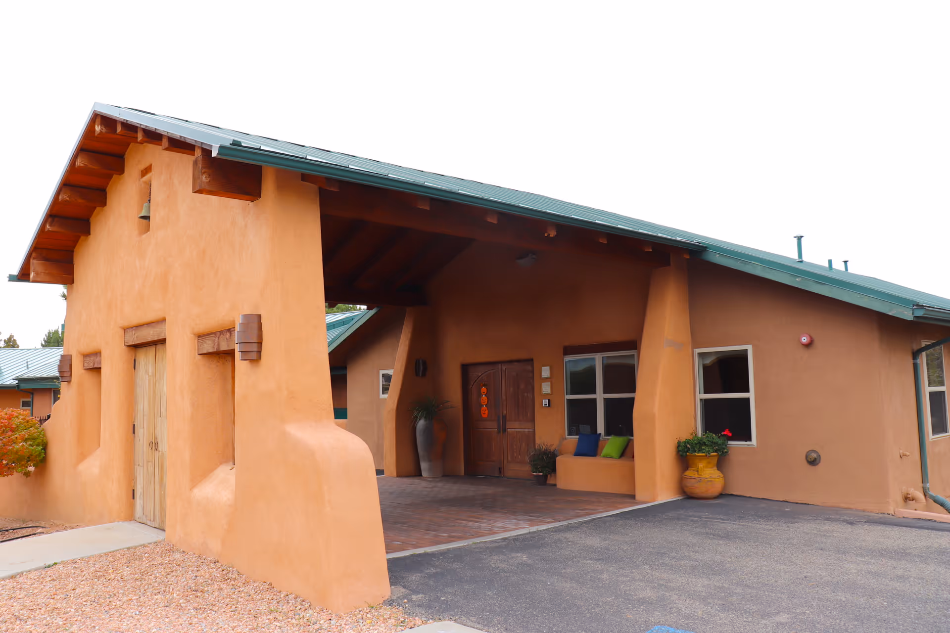 Exterior view of a building with adobe-style architecture, featuring a covered entrance with wooden double doors, a green metal roof, and potted plants near the entrance.