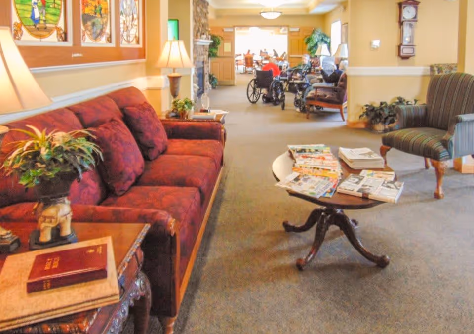 A cozy senior living facility lounge area with a red upholstered sofa, a wooden side table with a plant and books, a round wooden coffee table with magazines, and a striped armchair. In the background, several seniors are seated, some in wheelchairs, in a well-lit common area with warm yellow walls and decorative stained glass artwork.