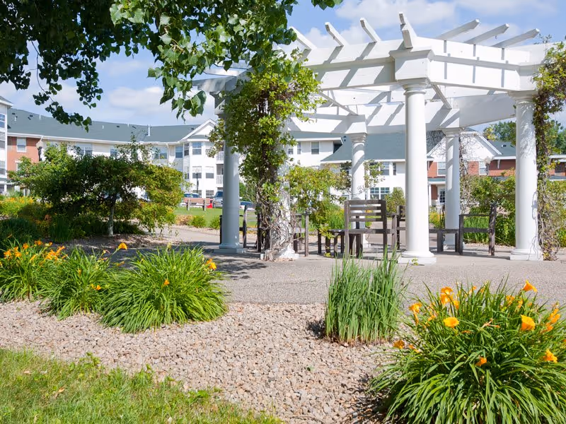 Outdoor garden area at The Rivers Retirement Community featuring a white pergola with climbing plants, several wooden chairs underneath, surrounded by green shrubs, flowering plants with orange blooms, and a gravel pathway. The retirement community building is visible in the background under a partly cloudy sky.