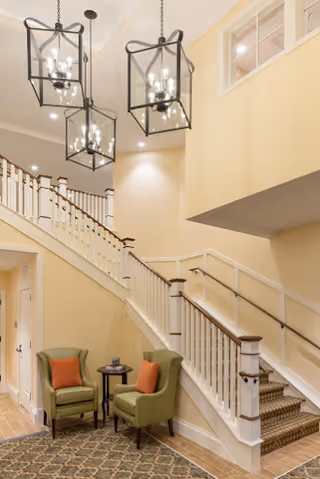 Bright interior lobby with a staircase, three hanging lantern chandeliers, and two green chairs beside a small table.