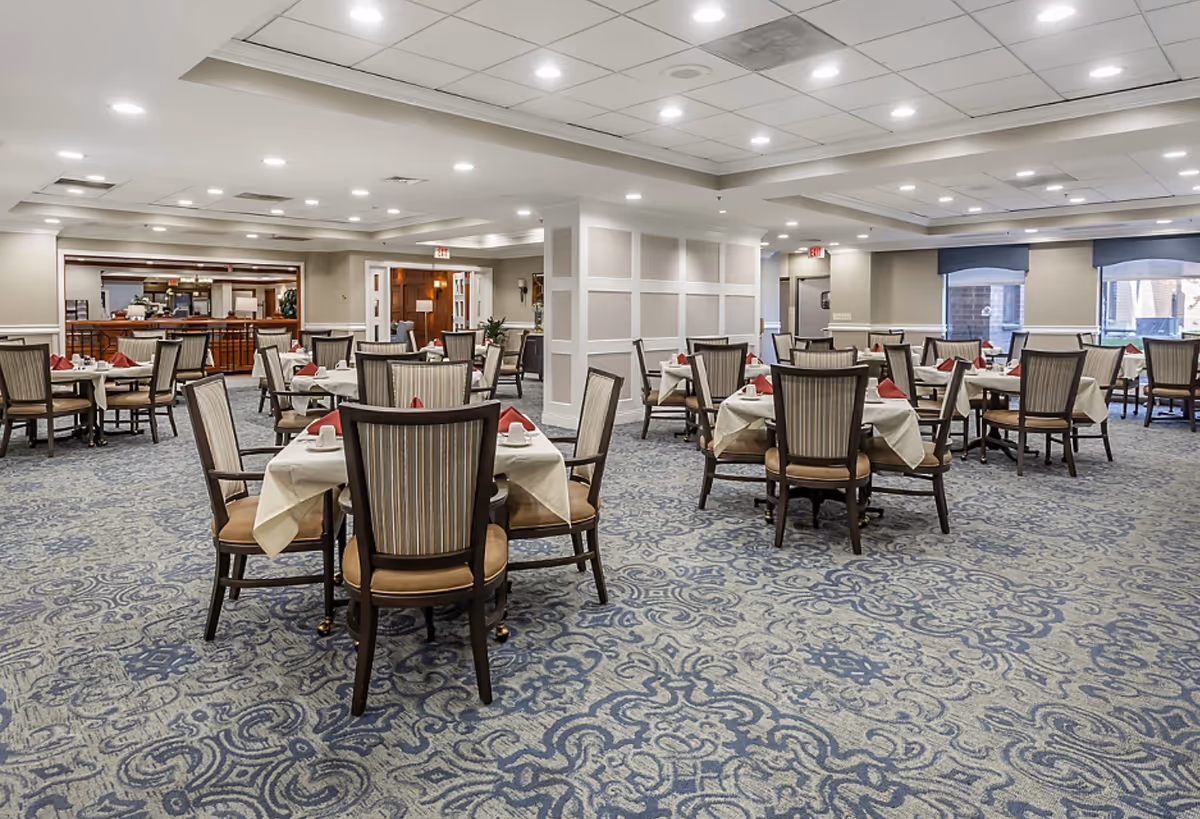 A spacious dining room with multiple round tables covered with white tablecloths and set with cups and red napkins. The room has patterned blue carpet flooring, beige walls, and large windows with blue valances allowing natural light to enter. The ceiling has recessed lighting and a coffered design.