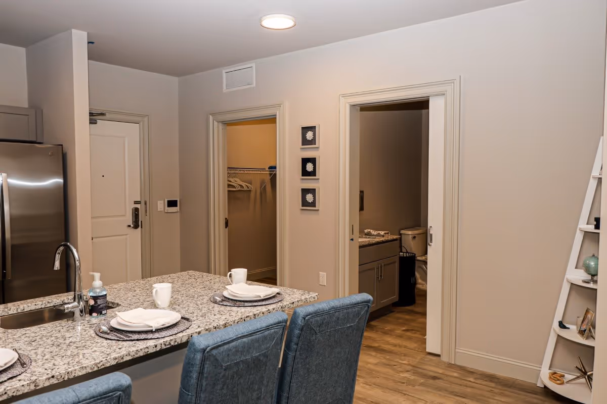 Interior view of a modern apartment kitchen area with a granite countertop island set with plates, cups, and napkins. Behind the island, there are two open doorways, one leading to a walk-in closet with hangers and the other to a bathroom with a visible toilet and vanity. The kitchen features stainless steel appliances and blue upholstered chairs at the island. A white ladder shelf with decorative items is visible on the right side.