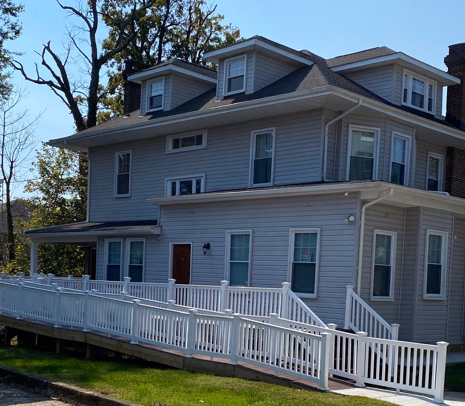 Three-story gray house with dormer windows and a white accessibility ramp and railing in front.