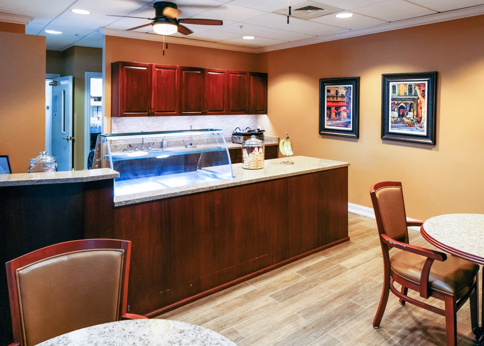 Interior view of a dining area with a serving counter featuring a glass sneeze guard, wooden cabinets, and a countertop with a jar of cookies and a banana holder. There are round tables with chairs and two framed paintings on the wall.