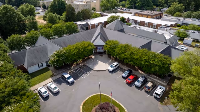 Aerial view of Rock Hill Post Acute Care Center showing a large building with a gray roof surrounded by trees and a parking lot with several cars parked. The entrance is visible with a covered walkway and a circular driveway with a flagpole in the center.