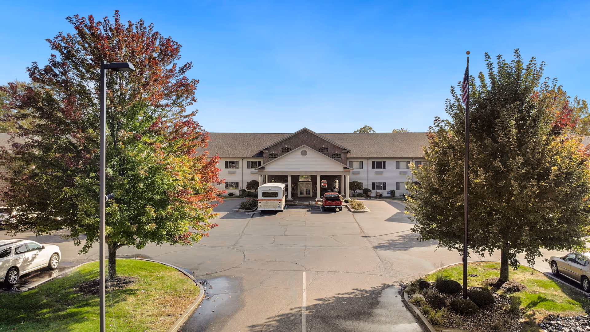 Front exterior view of a two-story senior living facility building with a covered entrance, surrounded by trees with autumn foliage, a parking lot with several cars, and a clear blue sky.