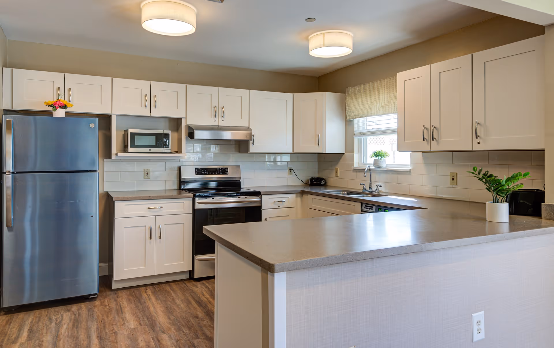 A modern kitchen with white cabinets, stainless steel refrigerator, stove, microwave, and dishwasher. The kitchen features a large countertop island, wood flooring, and a window with a beige valance letting in natural light. There are small potted plants on the countertop and windowsill.