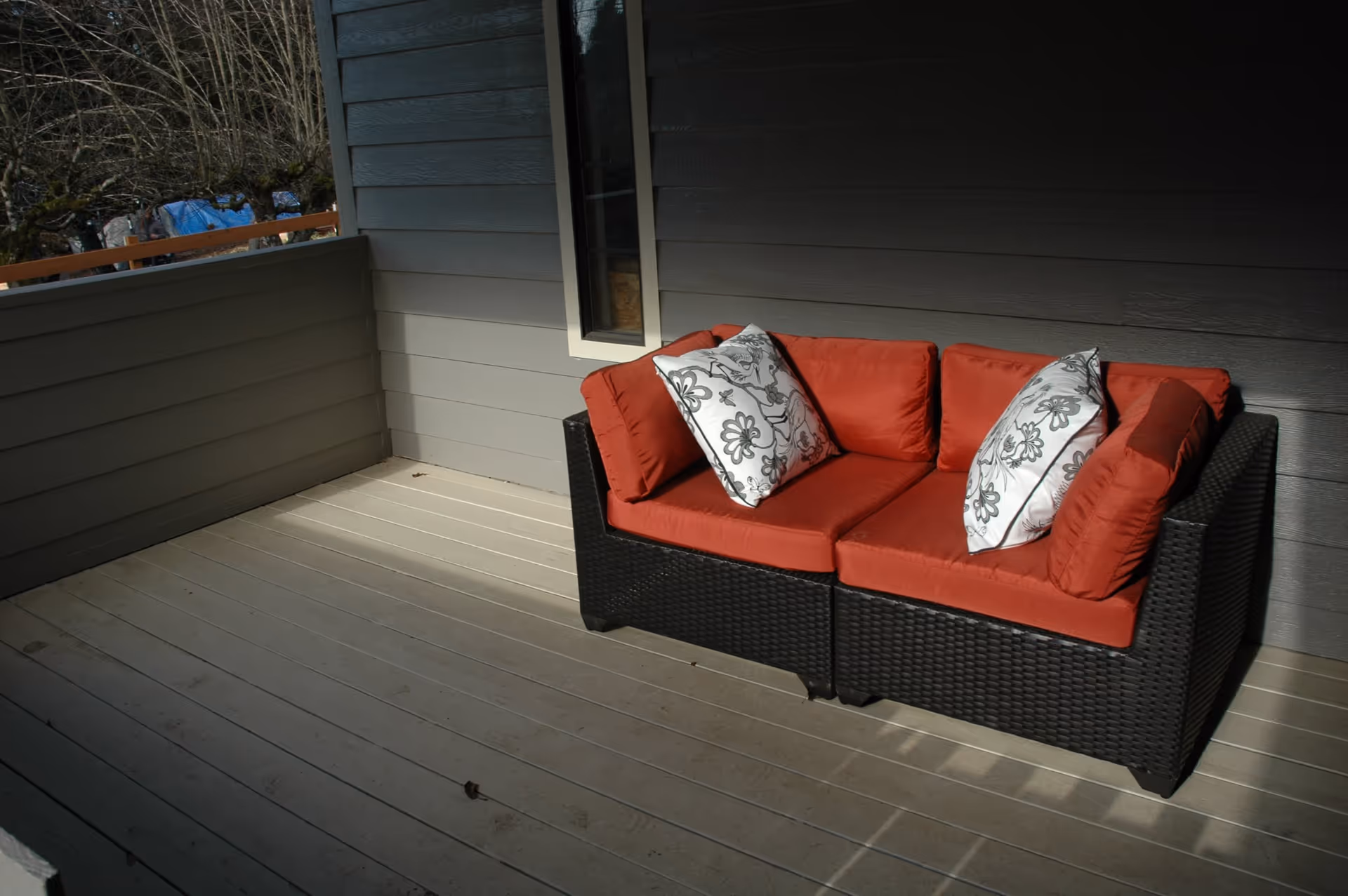 Outdoor patio area with a black wicker loveseat featuring red cushions and two white decorative pillows with floral patterns, against a gray wooden wall with a small vertical window.