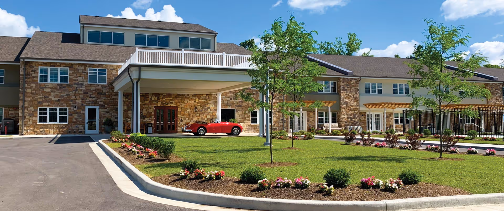 Front exterior of a senior living building with a covered entrance, landscaped lawn, and a red convertible parked under the porte-cochere.