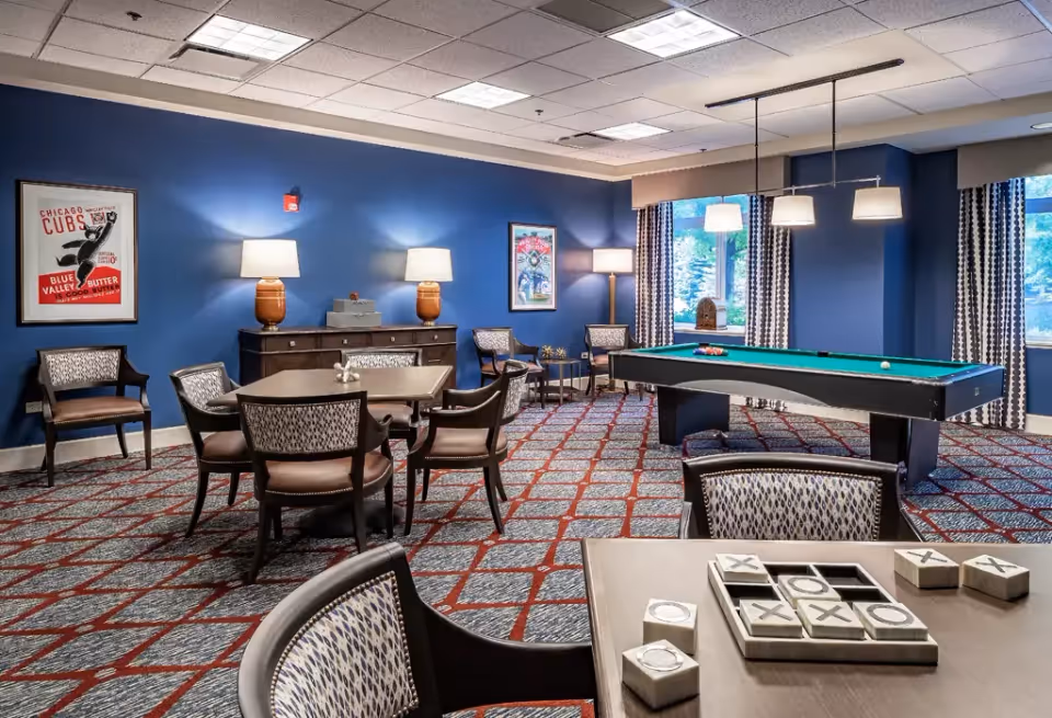 A recreational room with blue walls and patterned carpet featuring a pool table, several tables with chairs, and a tic-tac-toe game on the nearest table. The room is well-lit with ceiling lights and lamps, and has large windows with patterned curtains letting in natural light. There are framed posters on the walls and a wooden sideboard with lamps on it.