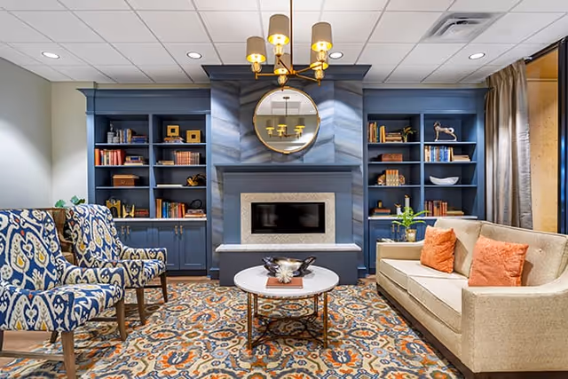 A cozy living room with a beige sofa adorned with orange pillows, two patterned armchairs, a round coffee table with decorative items, built-in blue bookshelves filled with books and decor, a modern fireplace, a round mirror above the fireplace, and a chandelier with five lights hanging from the ceiling.