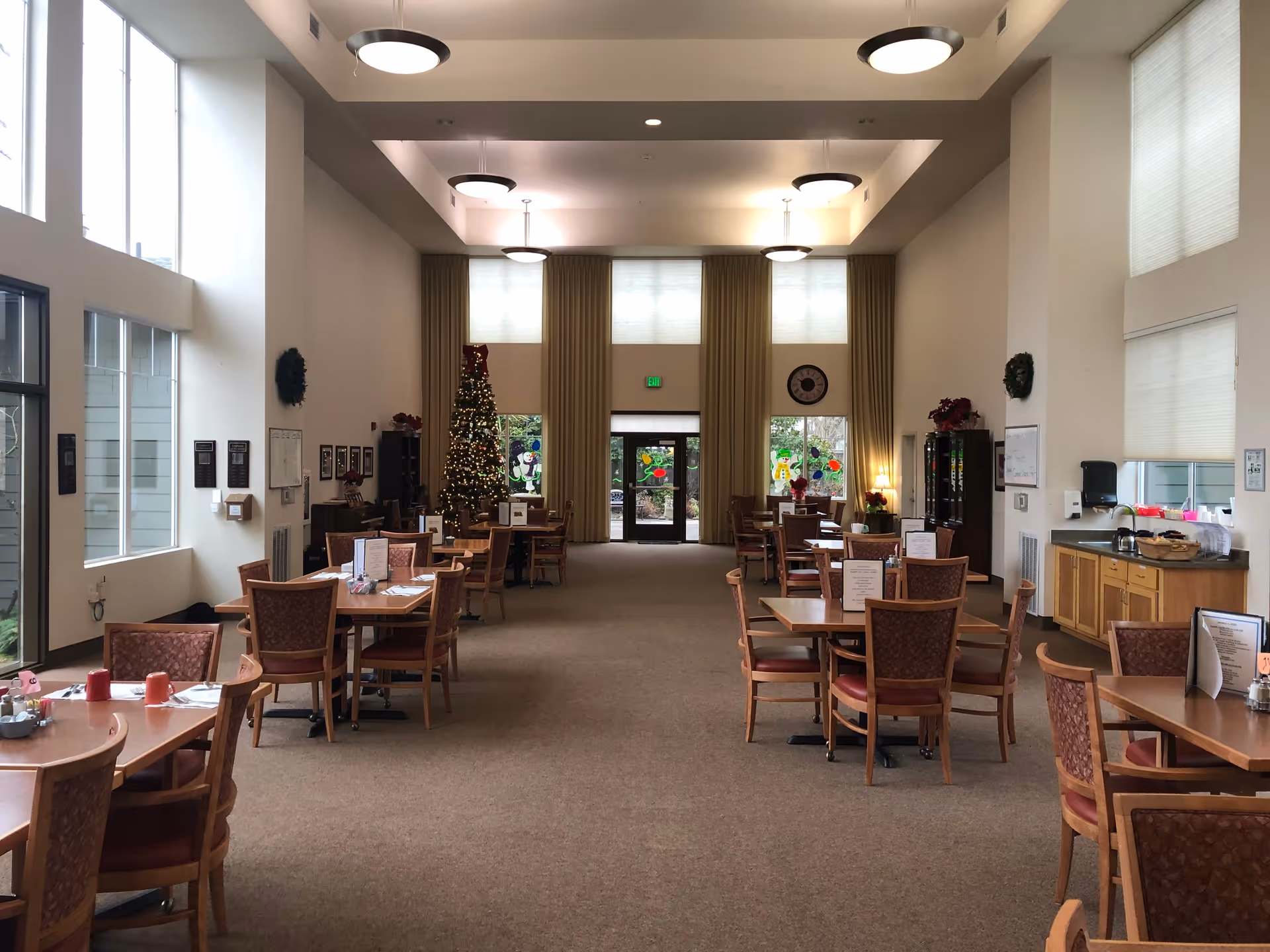 A spacious dining room with multiple wooden tables and chairs arranged neatly. The room has high ceilings with modern circular light fixtures. Large windows on the left side let in natural light. At the far end, there is a decorated Christmas tree and glass doors with holiday-themed window decorations. The walls are adorned with wreaths and framed pictures, and there is a counter with a coffee pot and supplies on the right side.