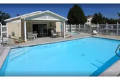 Outdoor swimming pool with clear blue water next to a small covered patio area with chairs and a table. The area is enclosed by a white fence, and there are trees and buildings visible in the background under a clear sky.