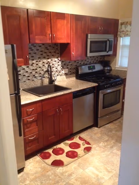 A compact kitchen with wooden cabinets, a stainless steel refrigerator, dishwasher, oven, and microwave. The backsplash features a mosaic tile design, and there is a black sink with a modern faucet. A small window with a floral valance is visible above the counter, and a beige floor mat with red apple designs is placed on the tiled floor.