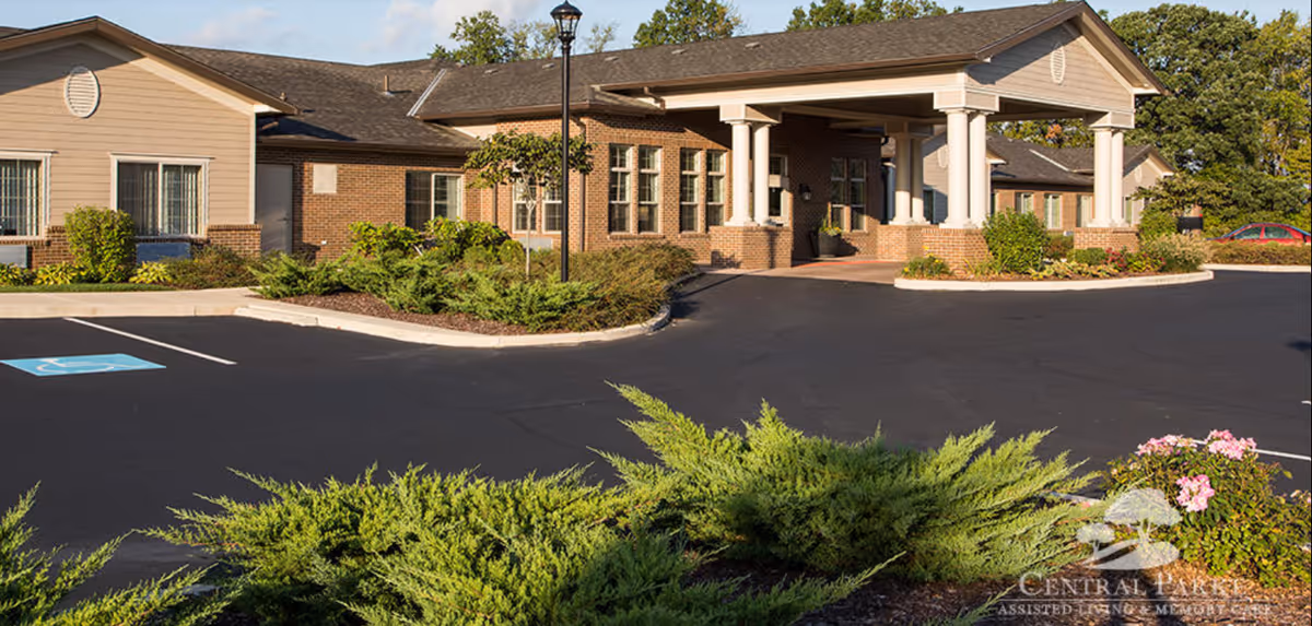 Exterior view of Central Parke Assisted Living & Memory Care building with a covered entrance, surrounded by greenery and a parking lot with a handicapped parking space.