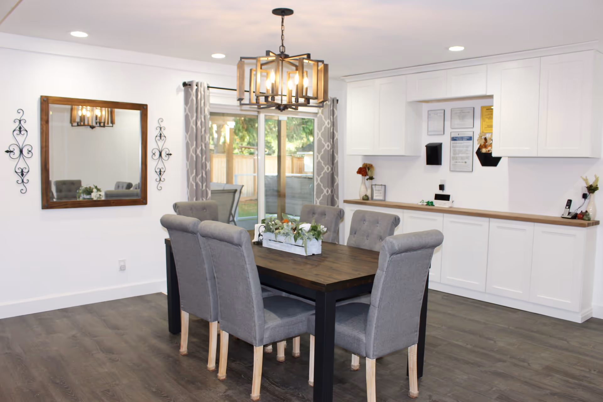 A dining room with a dark wooden table surrounded by six gray upholstered chairs. A rectangular chandelier with exposed bulbs hangs above the table. On the table is a white planter box with green plants. The room has white walls, a large mirror with a wooden frame on one wall, and two decorative black metal wall hangings. There is a sliding glass door with gray patterned curtains leading outside. White cabinets with a wooden countertop line one wall, with some framed documents and small decorative items on the counter.
