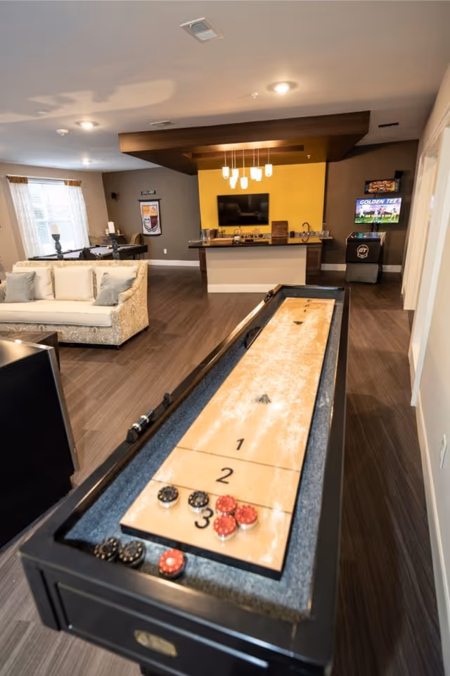 A lounge-style common room with a shuffleboard table in the foreground, sofas, a TV, and a small bar area in the background.