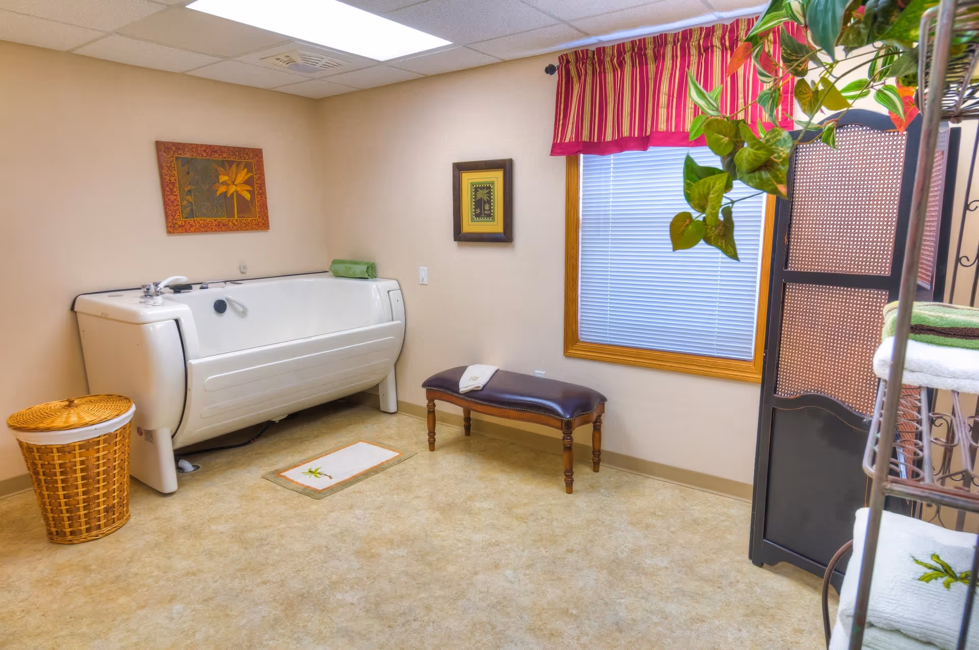 A clean and well-lit bathing room featuring a large white walk-in bathtub, a wicker laundry basket, a small bench with a folded towel, a window with blinds and a red striped valance, two framed pictures on the wall, and a plant hanging near a black folding screen.