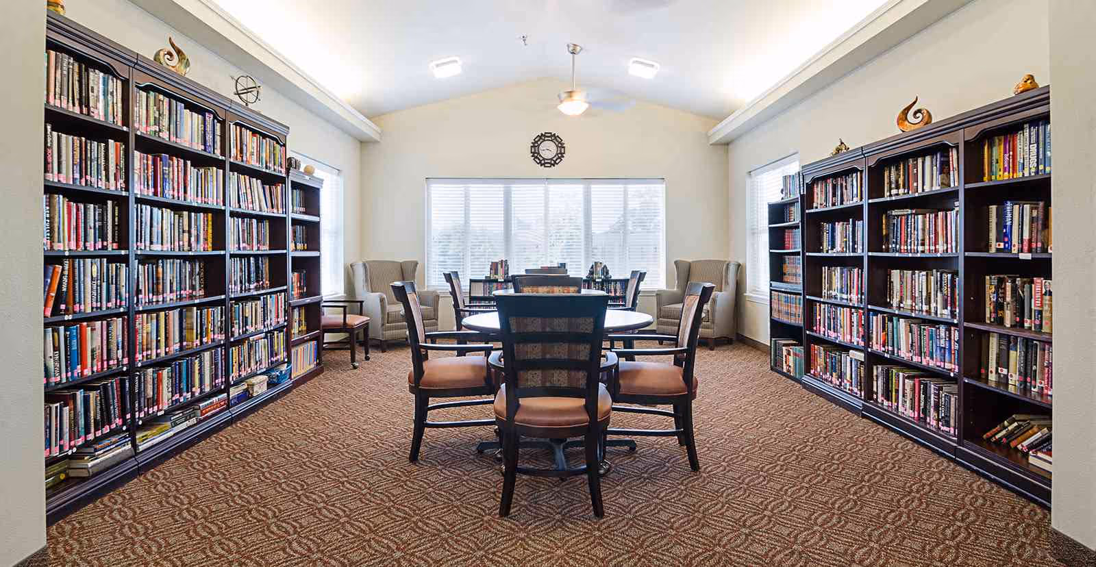 A bright and spacious library room with two large dark wood bookshelves filled with books on either side. In the center, there is a round table surrounded by six chairs with cushioned seats. At the back of the room, two armchairs are positioned near a large window with blinds, and a wall clock is mounted above the window. The room has a patterned carpet and a ceiling fan with lights.