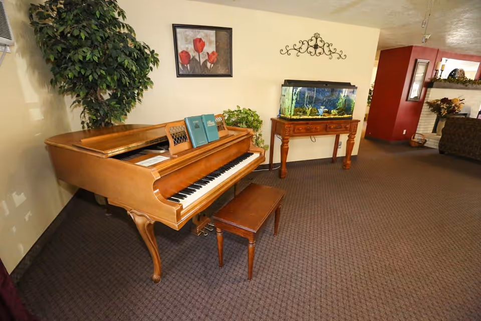 A cozy interior room featuring a wooden grand piano with an open lid and a matching bench. On the piano, there are two music books. To the right, there is a wooden table holding a fish tank with aquatic plants and decorations. The room has beige walls adorned with a framed picture of red flowers and a decorative metal wall piece. A large green potted plant is placed in the corner near the piano. The carpet is brown with a subtle pattern, and part of a fireplace and seating area is visible in the background.