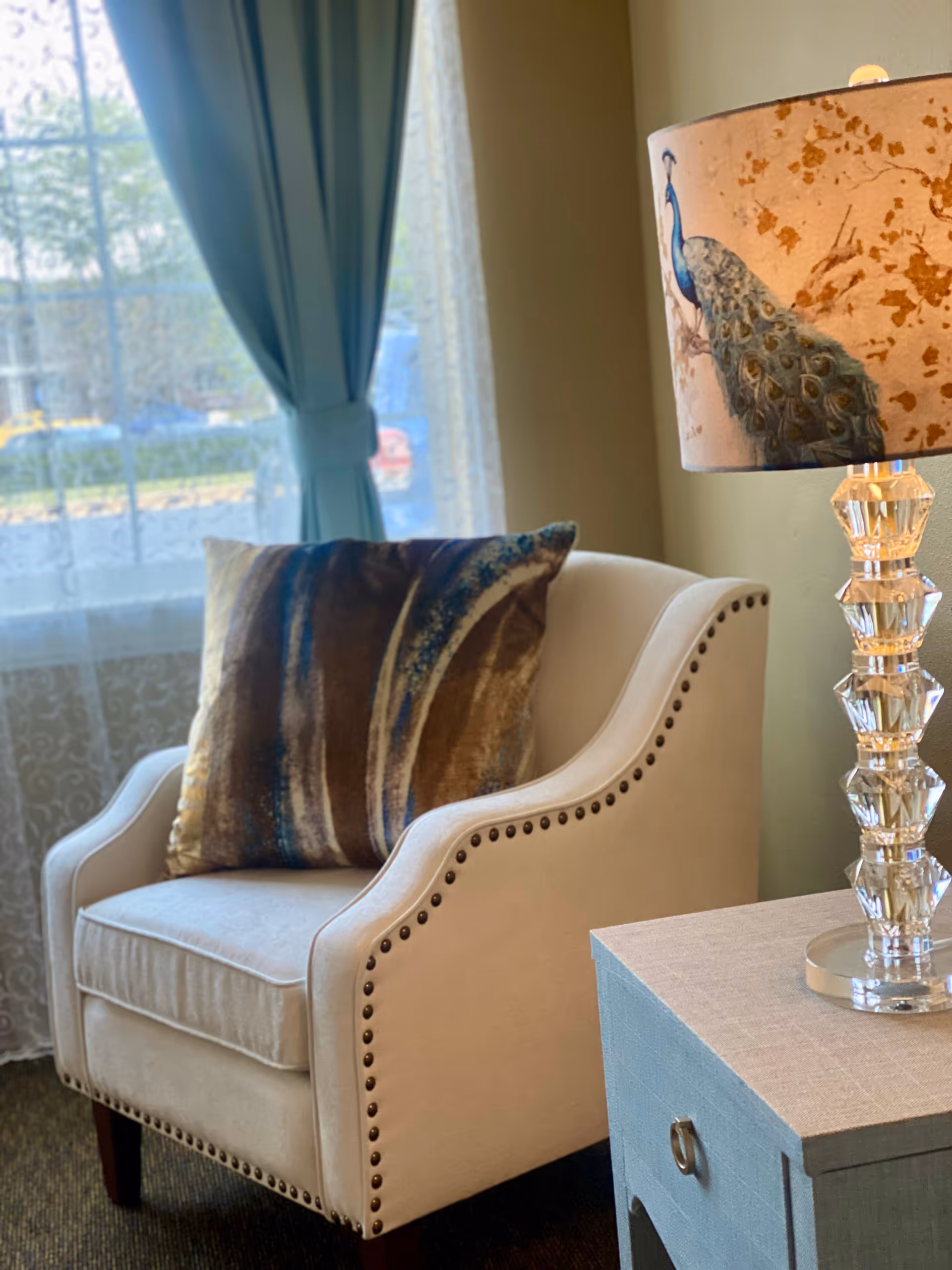 A beige upholstered armchair with nailhead trim and a decorative pillow with brown and blue patterns is positioned next to a light blue side table. On the table is a lamp with a clear, faceted base and a lampshade featuring a peacock design. Behind the chair is a window with sheer white curtains and blue drapes tied back, allowing natural light to enter the room.