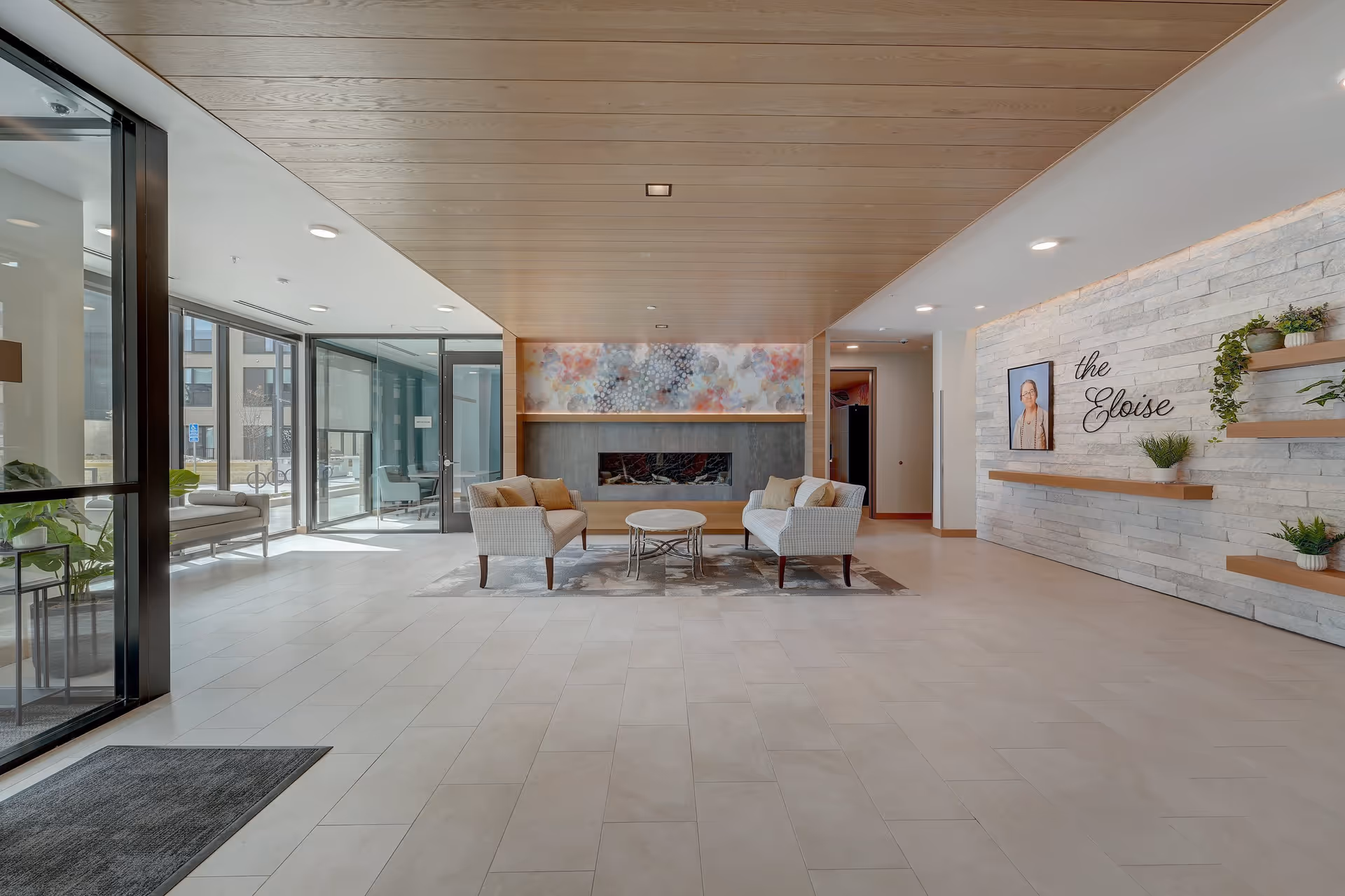 A modern and spacious lobby area with a light wood-paneled ceiling and light tiled floor. Two cushioned armchairs and a small round table are arranged on a rug in front of a decorative fireplace with colorful artwork above it. To the right, a stone accent wall features the words 'the Eloise' in cursive, a framed portrait, and several shelves with potted plants. Large glass windows and doors on the left allow natural light to fill the space.