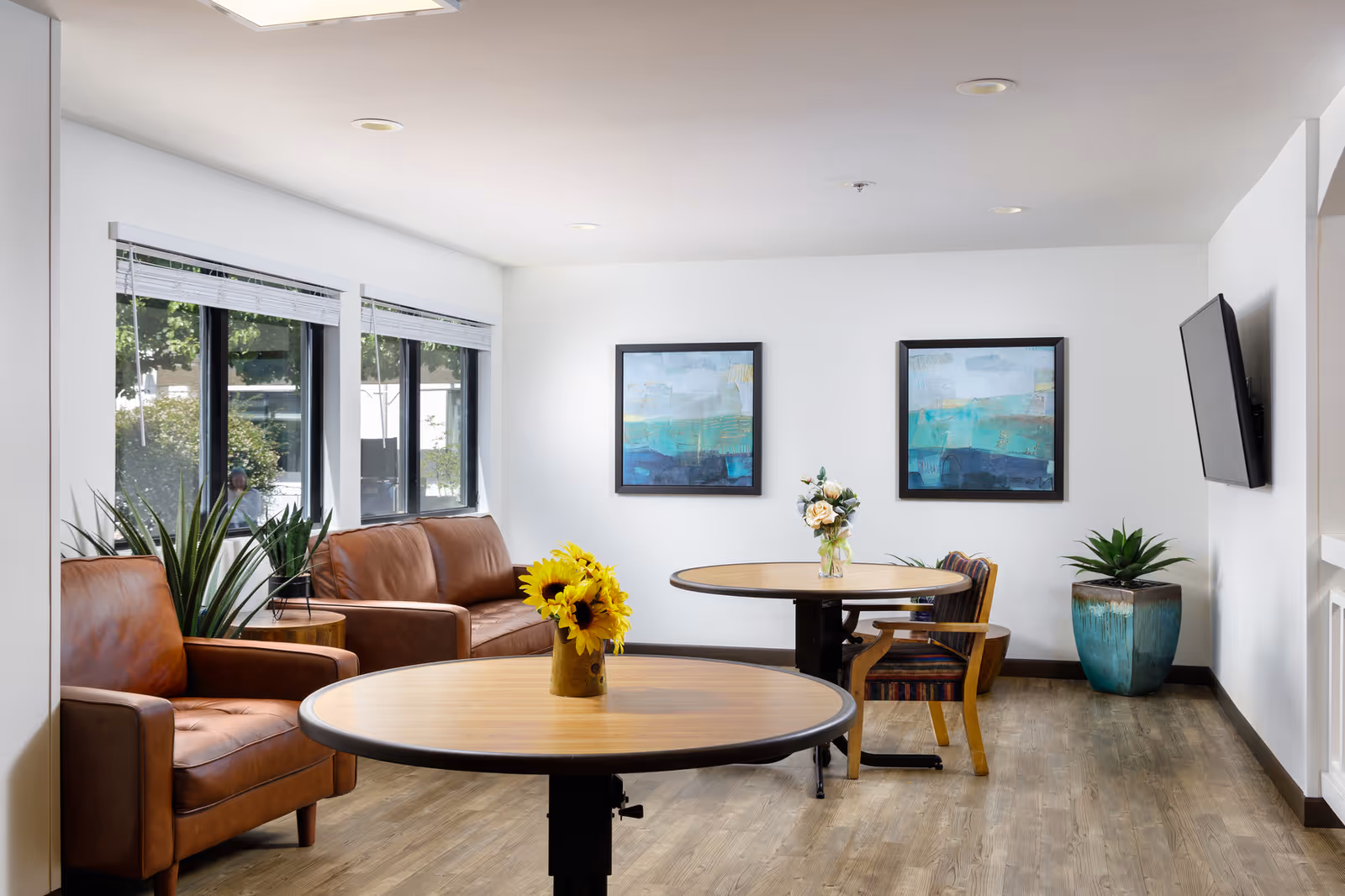 A bright and clean common area with two round wooden tables, each decorated with a vase of flowers. There are two brown leather armchairs and a matching sofa near large windows with white blinds. The walls are white and adorned with two abstract paintings. A flat-screen TV is mounted on the right wall, and there are green potted plants placed around the room on a wood floor.