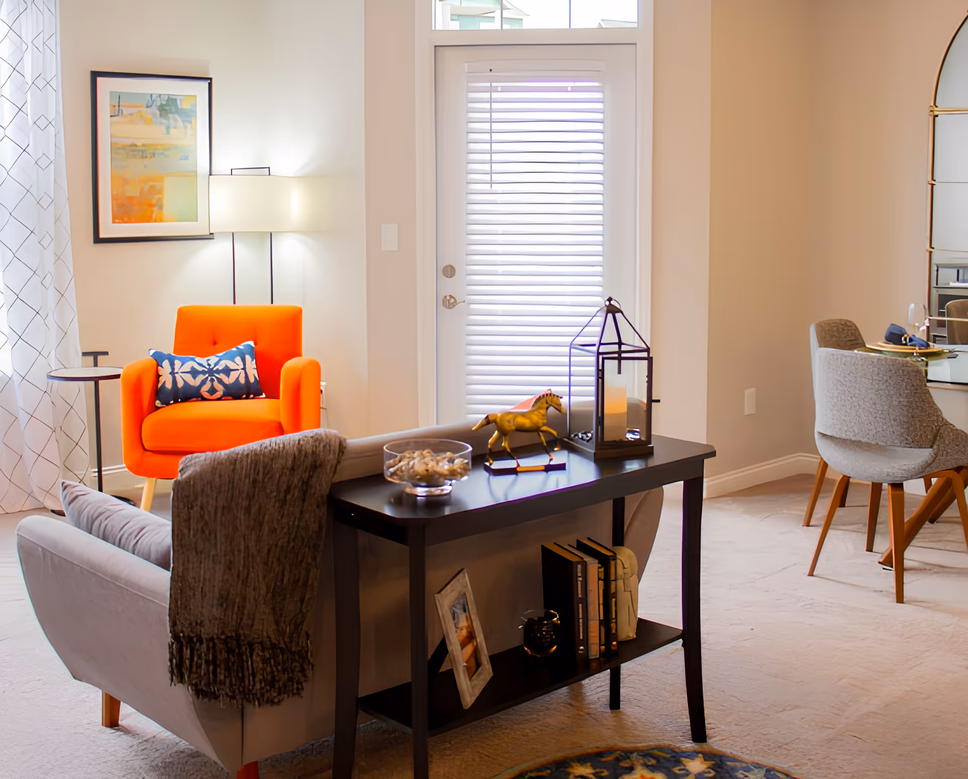 A cozy living room area with a bright orange armchair featuring a blue patterned pillow, a gray sofa with a dark throw blanket, and a black console table decorated with books, a framed photo, a decorative horse figurine, and a lantern. A floor lamp and framed artwork are on the wall behind the armchair, and a dining area with gray chairs and a large mirror is visible to the right.