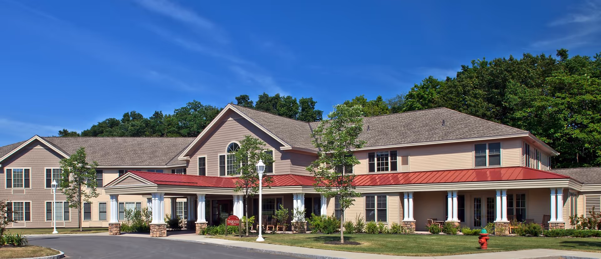 Two-story beige senior living building with a red metal awning, white columns at the entrance, landscaped lawn and trees under a blue sky.
