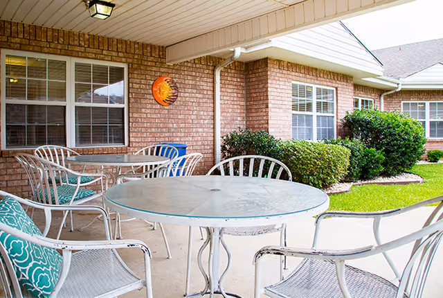 Covered outdoor patio area with white metal tables and chairs, some with green patterned cushions, adjacent to a brick building with windows and green bushes along the side.