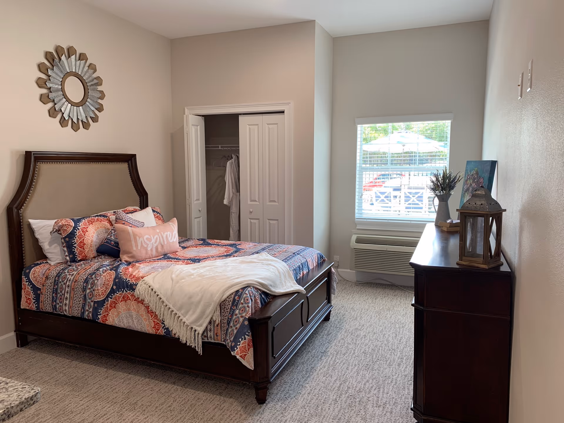 A cozy bedroom with a wooden bed frame featuring a colorful patterned bedspread and multiple pillows, including one with the word 'Inspired'. There is a white blanket draped over the foot of the bed. A closet with double doors is open, showing hanging robes inside. A window with blinds lets in natural light, and below it is a wall-mounted air conditioning unit. A dark wooden dresser with decorative items, including a lantern and a vase with flowers, is positioned against the right wall. The room has beige walls and carpeted flooring.
