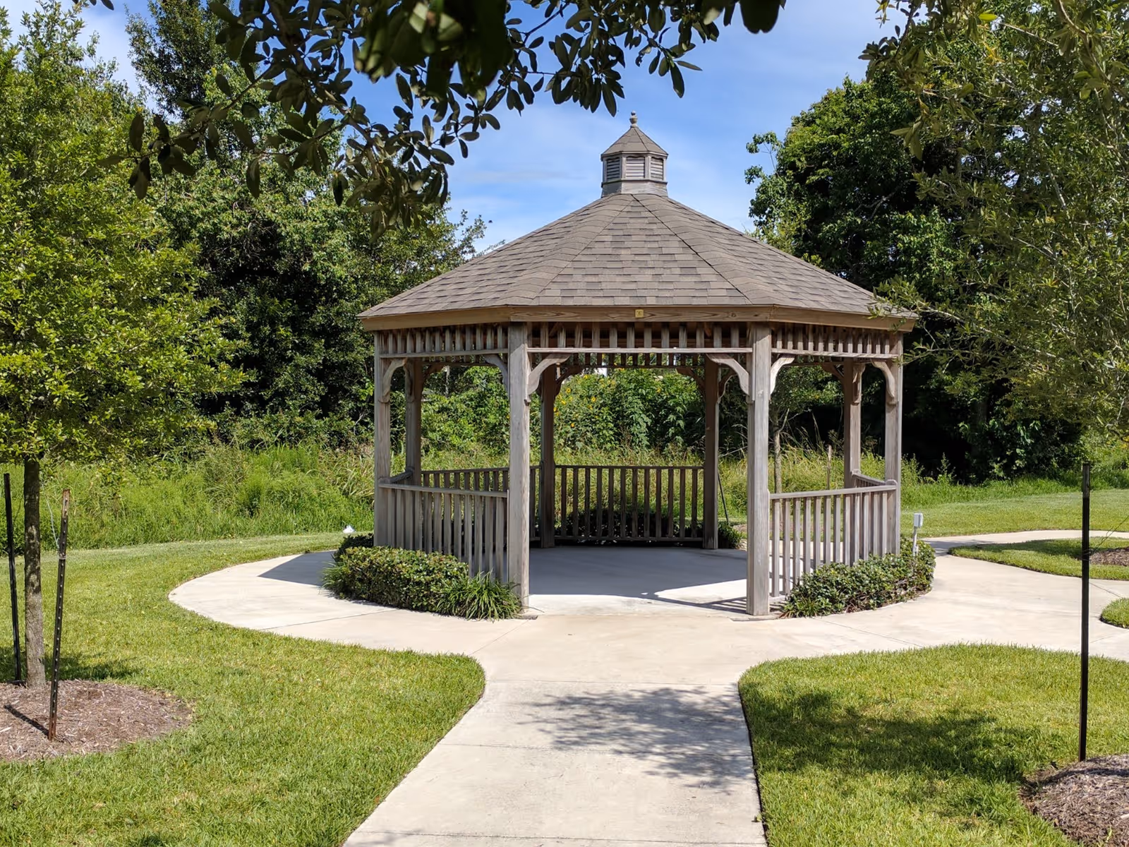 Wooden octagonal gazebo on a circular concrete path surrounded by grass and trees