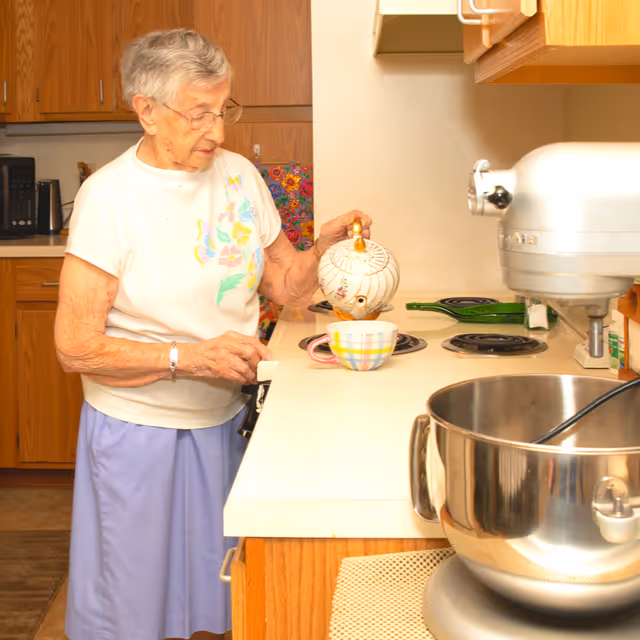 An elderly woman in a kitchen pouring tea from a decorative teapot into a mug on the counter.